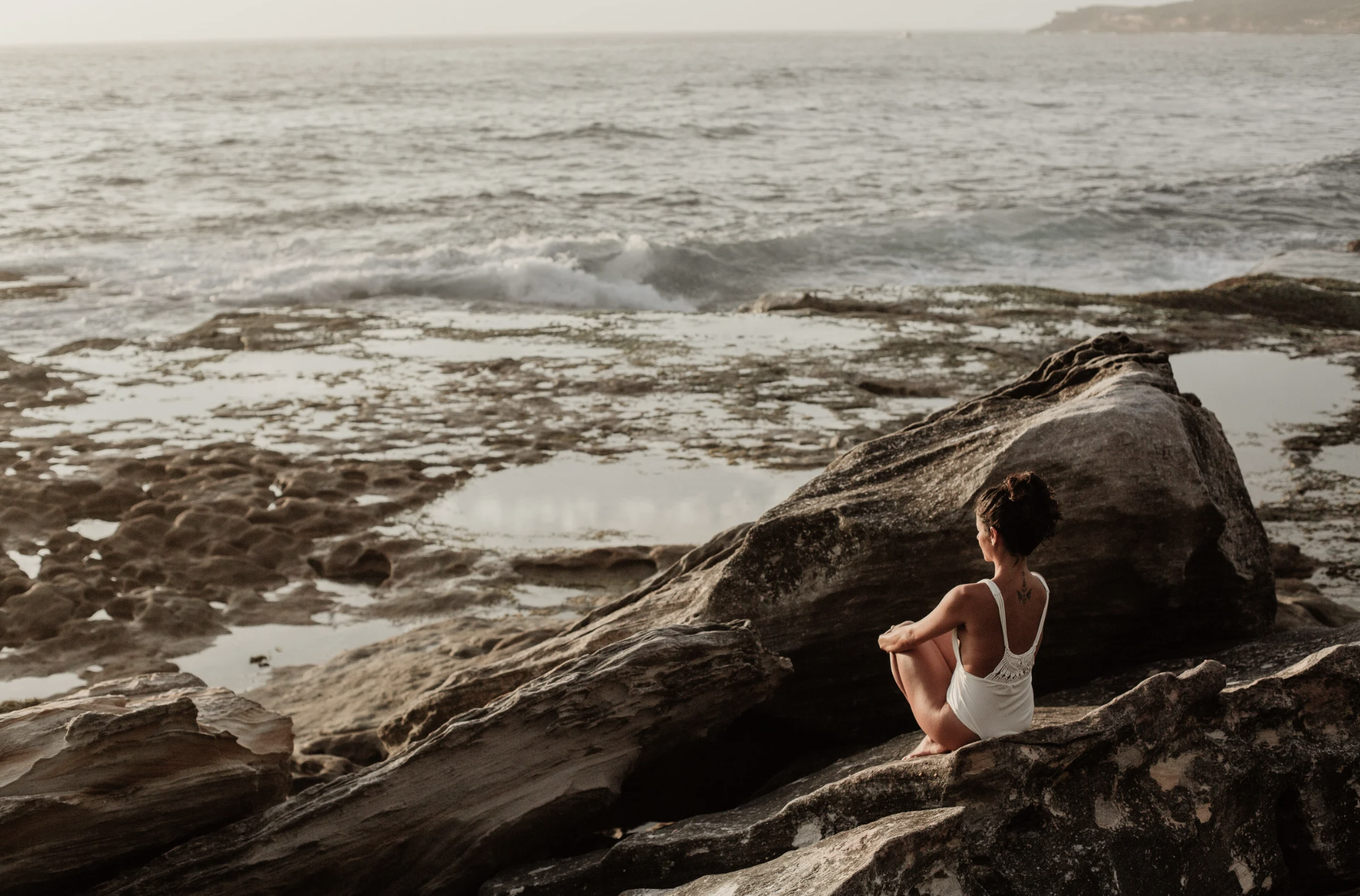 woman sitting on a rocky shoreline looking at the ocean waves.